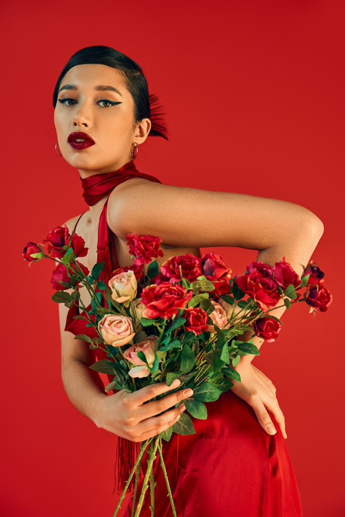 jeune et séduisante femme asiatique avec un maquillage audacieux, en robe à la mode posant avec la main sur la hanche et les roses tout en regardant la caméra sur fond rouge, printemps à la mode, tournage de mode - Photo, image