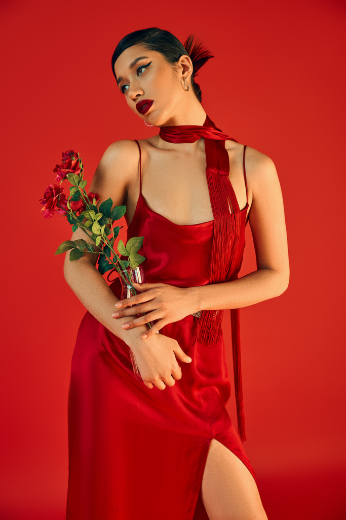модная весна, юная мода, привлекательная азиатка с брюнетками и смелым макияжем, в элегантном платье с ремешком и шейным платком, держащая стеклянную вазу со свежими розами на красном фоне - Фото, изображение