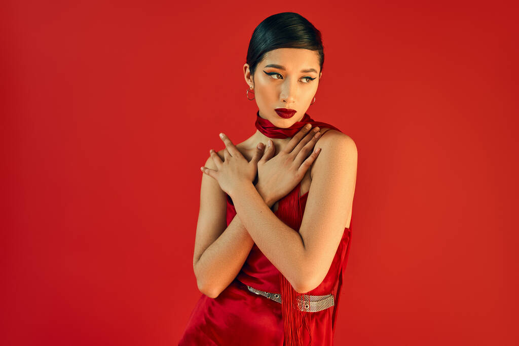 young asian woman with brunette hair and bold makeup holding hands on chest and looking away while posing in neckerchief and dress on red background, fashionable spring - Photo, Image