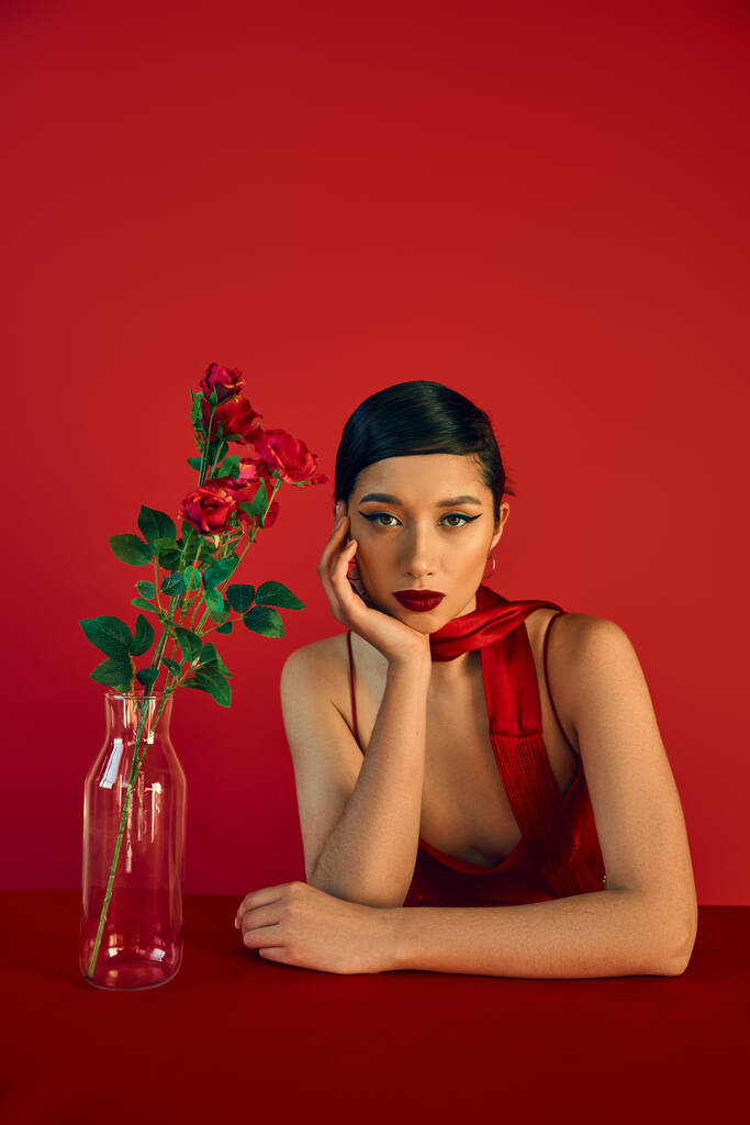 μοντέρνα άνοιξη, νεανική μόδα, όμορφη Ασιάτισσα γυναίκα με τολμηρό μακιγιάζ και καστανά μαλλιά κάθεται στο μαντήλι κοντά σε γυάλινο βάζο με τριαντάφυλλα και κοιτάζοντας κάμερα σε κόκκινο φόντο - Φωτογραφία, εικόνα