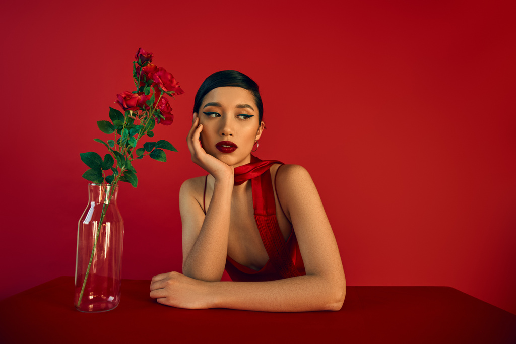 alluring asian woman with brunette hair and bold makeup, in elegant dress and neckerchief sitting at table near glass vase with fresh roses on red background, spring fashion concept - Photo, Image