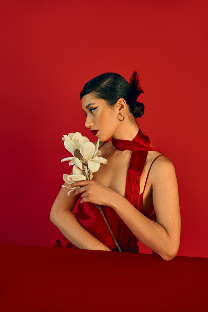 concetto di moda primaverile, giovane donna asiatica seduta a tavola con orchidea bianca e guardando da un'altra parte su sfondo rosso, capelli castani, trucco audace, cinturino, fazzoletto, gen z style - Foto, immagini