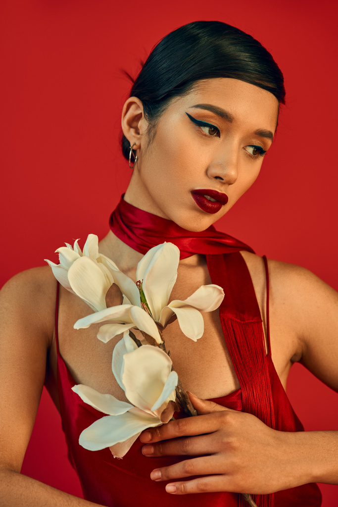 portré gyönyörű ázsiai nő elegáns pánt ruha, nyakkendő, merész smink és barna haj gazdaság fehér virágzó orchidea piros háttér, gen z divat, stílusos tavaszi koncepció - Fotó, kép