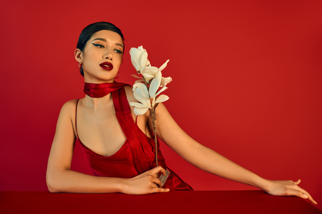 mujer asiática elegante y encantadora en vestido elegante y pañuelo, con pelo moreno y maquillaje audaz mirando la orquídea floreciente cerca de la mesa sobre fondo rojo, fotografía de moda de primavera - Foto, imagen