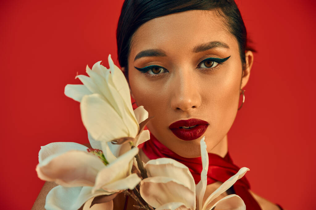 portrait of youthful and mesmerizing asian woman with brunette hair, bold makeup and expressive gaze looking at camera near while blooming orchid on red background, spring fashion photography - Photo, Image