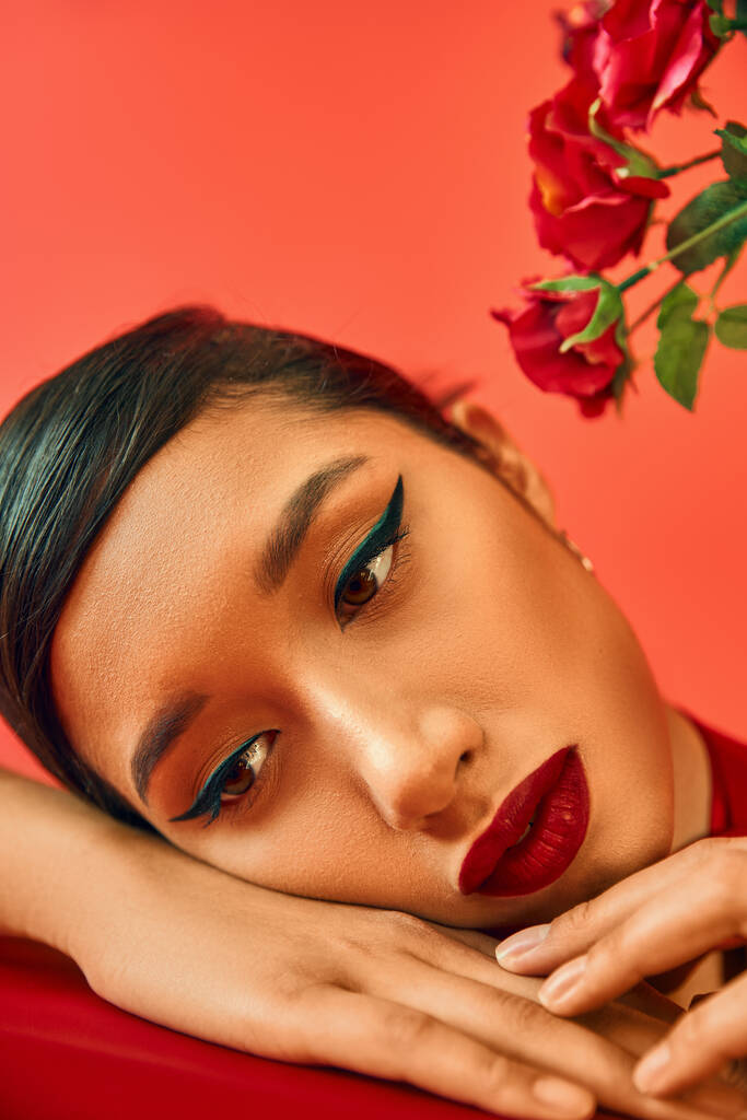 close up portrait of mesmerizing and pensive asian woman with bright makeup and brunette hair dreaming with hands near face next to flowers on red background, spring style, generation z - Photo, Image