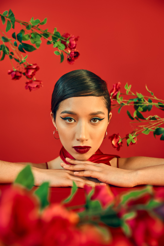 retrato de mujer asiática joven y encantadora con maquillaje brillante y cabello moreno mirando a la cámara rodeada de rosas frescas sobre fondo rojo, concepto de fotografía de moda de primavera - Foto, Imagen