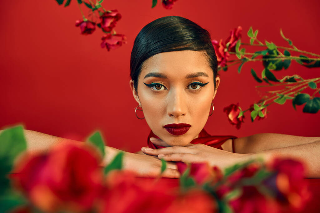 gen z mody, portret uroczej i młodej azjatyckiej kobiety z jasnym makijażem patrząc na aparat fotograficzny wśród świeżych róż na czerwonym, wiosenna fotografia mody, rozmyty pierwszy plan - Zdjęcie, obraz