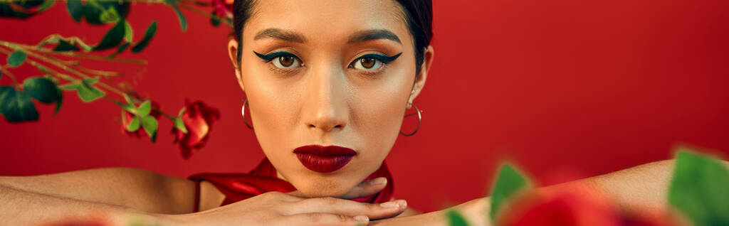 portrét mladistvého a okouzlujícího asijského módního modelu s výrazným make-upem a expresivním pohledem na kameru poblíž květin na červeném pozadí, trendy jarní koncept, banner - Fotografie, Obrázek