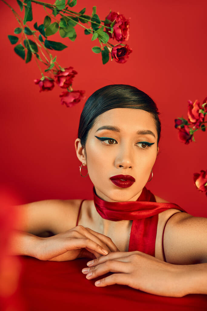 portrait of mesmerizing and dreamy asian woman posing near fresh roses on red background, bold makeup, brunette hair, stylish neckerchief, trendy spring concept - Photo, Image