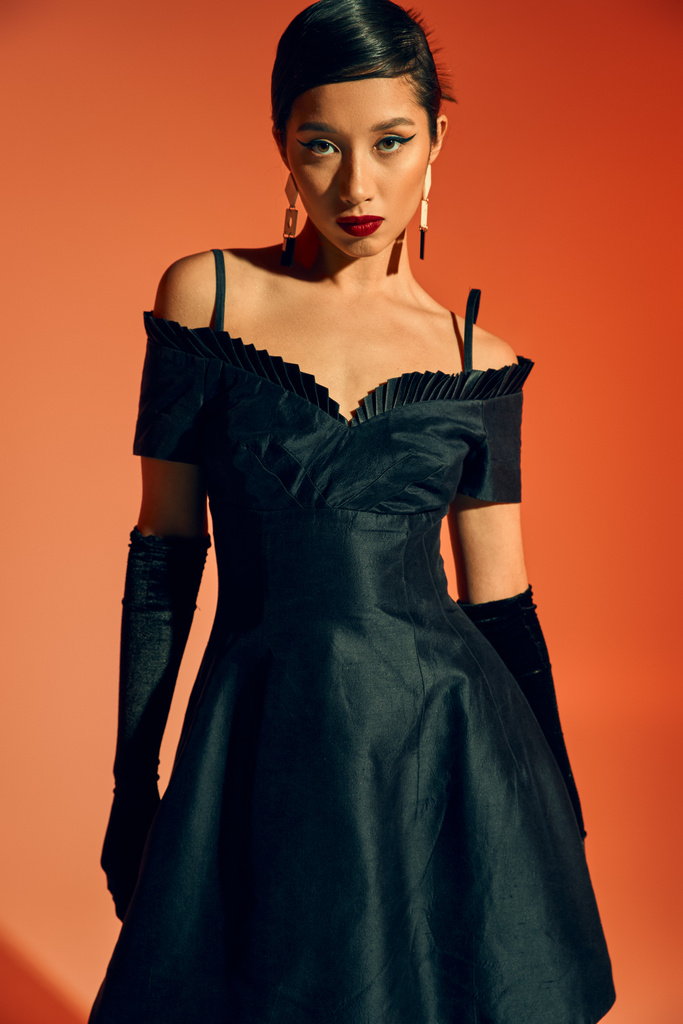 full length of fashionable and young asian woman in gloves, black cocktail dress and trendy earrings looking at camera with seductive gaze on red and orange background - Photo, Image