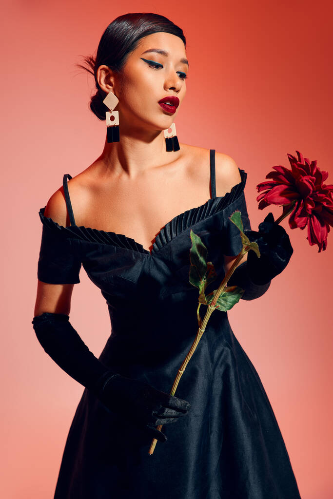 mujer asiática joven y de moda con maquillaje audaz, en guantes largos negros y vestido de cóctel mirando peonía borgoña mientras está de pie sobre fondo rojo y rosa, fotografía de moda de primavera - Foto, imagen