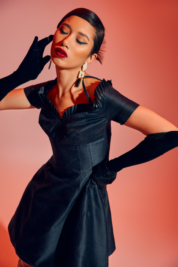 завораживающая азиатская женщина с смелым макияжем, брюнетками, в длинных перчатках и черном коктейльном платье, стоящая в стильной позе с закрытыми глазами на красном и розовом фоне, весенняя концепция моды - Фото, изображение