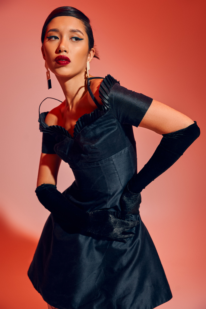 εκφραστική και μοντέρνα ασιατική γυναίκα σε μοντέρνα σκουλαρίκια, μακριά γάντια και μαύρο φόρεμα κοκτέιλ στέκεται με τα χέρια στο ισχίο και κοιτάζοντας μακριά σε κόκκινο και ροζ φόντο, μόδα άνοιξη, γενιά z - Φωτογραφία, εικόνα