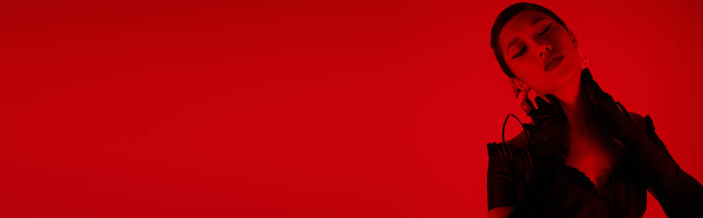 verleidelijke aziatische vrouw in zwart elegante jurk en lange handschoenen aanraken van de nek, terwijl poseren met gesloten ogen op levendige achtergrond met rode verlichting effect, jeugd cultuur, stijlvolle lente concept, banner - Foto, afbeelding