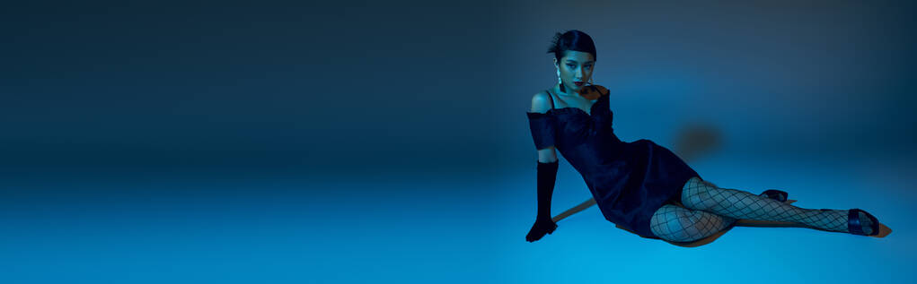full length of expressive asian woman in elegant cocktail dress, black long gloves and fishnet tights sitting on blue background with cyan lighting effect, generation z, spring style concept, banner - Photo, Image