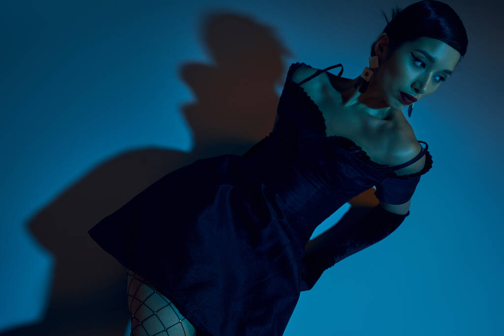 vista de alto ângulo de atraente mulher asiática com cabelo morena, em vestido de cocktail preto e luvas longas olhando para longe enquanto sentado no fundo azul com efeito de iluminação ciano - Foto, Imagem