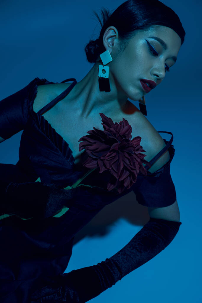 γοητευτική Ασιάτισσα γυναίκα σε μαύρο κομψό φόρεμα, μακριά γάντια και μοντέρνα σκουλαρίκια κάθεται σε κομψή στάση με παιώνι λουλούδι σε μπλε φόντο με κυανό φωτισμό αποτέλεσμα, μοντέρνα έννοια άνοιξη - Φωτογραφία, εικόνα