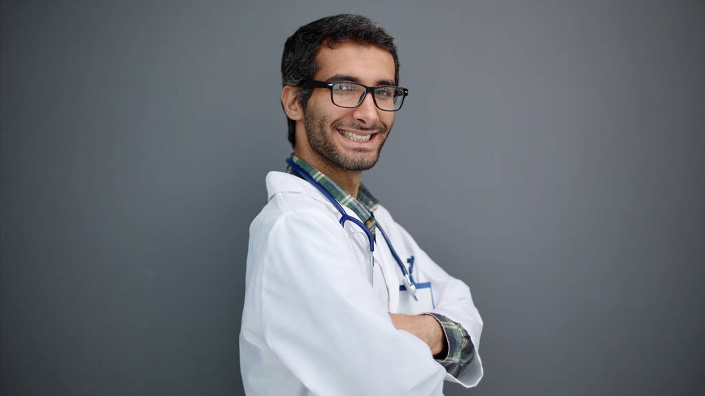 Portret van een positieve mannelijke arts poseren met gekruiste armen en glimlachen op camera over grijze studio achtergrond - Foto, afbeelding
