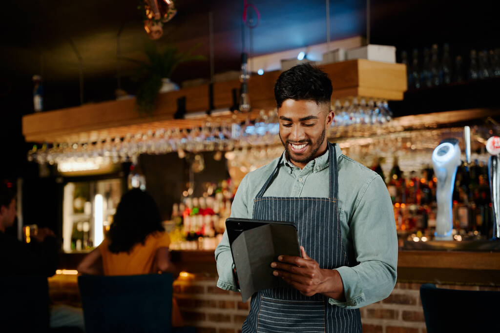 Happy young biracial man wearing apron using digital tablet while working as waiter in restaurant - Photo, Image
