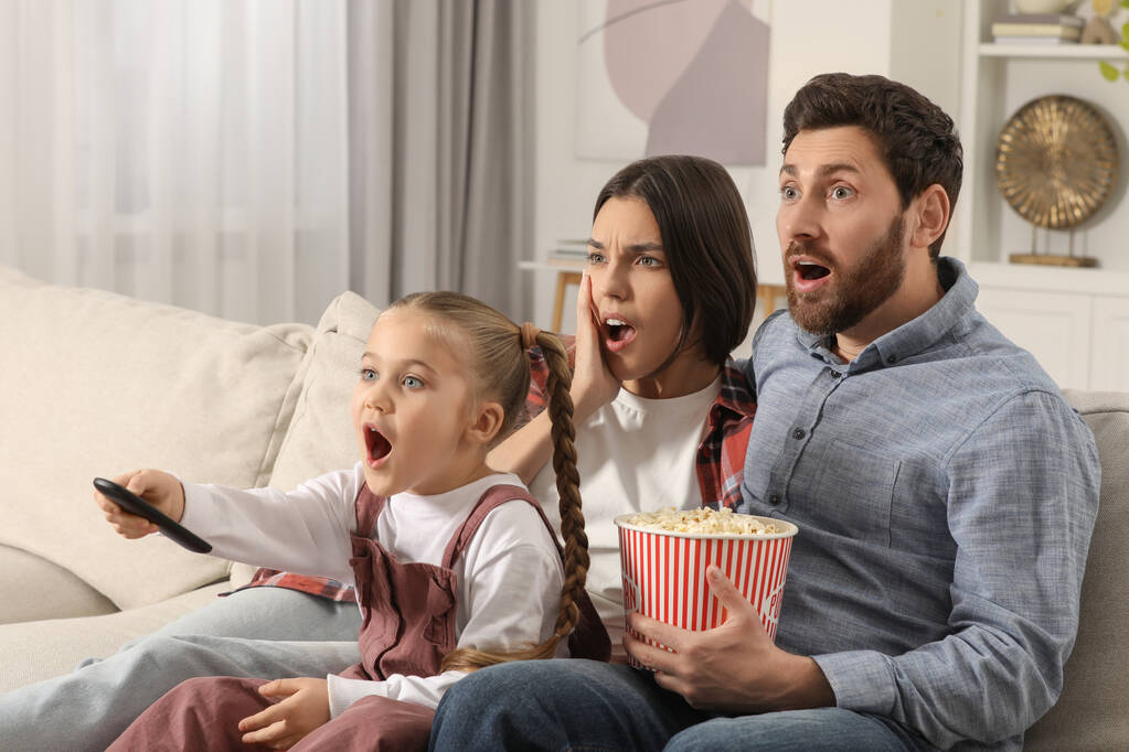 Удивлённая семья смотрит телевизор с попкорном на диване дома - Фото, изображение