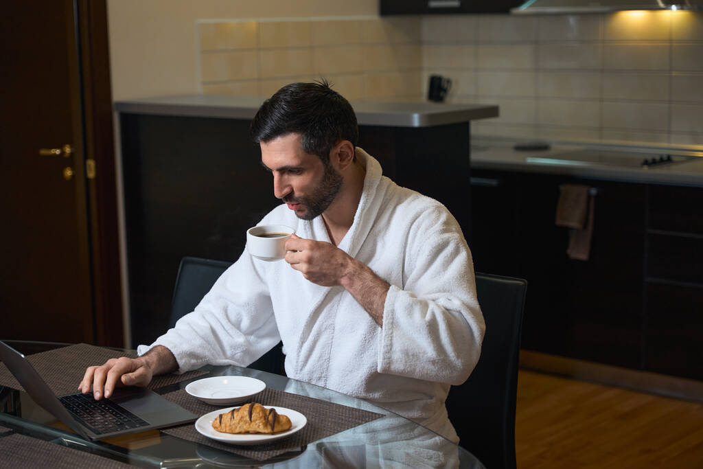 L'homme s'assoit avec un café du matin devant un ordinateur portable dans la cuisine et la salle à manger - Photo, image