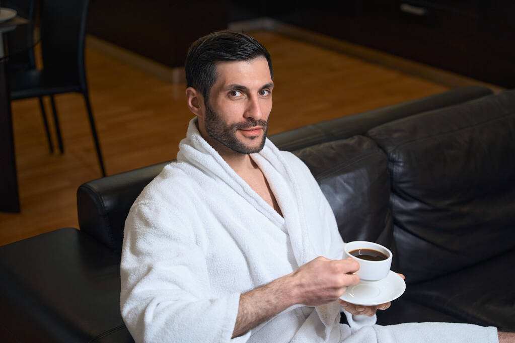 Hotelový host středního věku si vychutnává šálek ranní kávy, muž sedí na pohodlné kožené pohovce - Fotografie, Obrázek