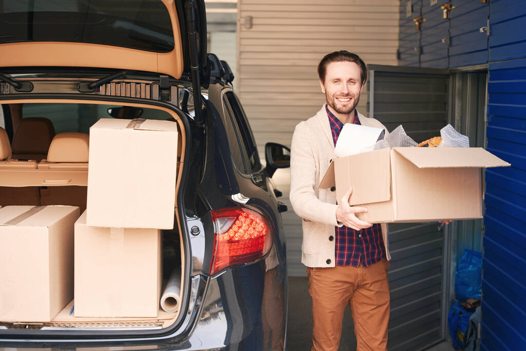 Uomo felice è in piedi vicino al bagagliaio aperto di una macchina con scatole in un magazzino e in possesso di una grande scatola di cartone - Foto, immagini