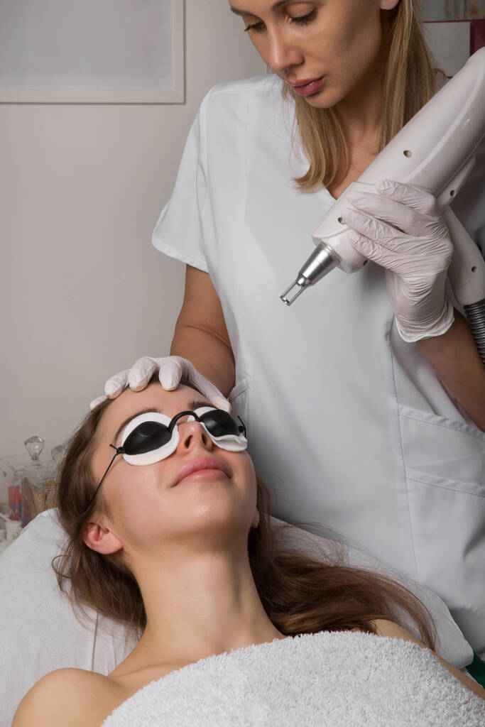 schoonheidsspecialiste met beauty laser in de buurt van het gezicht van de vrouw, beschermende brillen, spa procedures met laser en zwarte maskers. Hoge kwaliteit foto - Foto, afbeelding