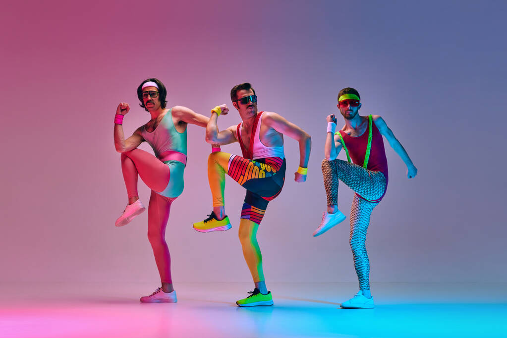 Sport show. Trzech zabawnych mężczyzn w kolorowych strojach sportowych wykonujących ćwiczenia aerobiku na tle gradientowego niebieskiego różowego studia w neonowym świetle. Koncepcja sportowego i aktywnego stylu życia, humoru, stylu retro. Ogłoszenie - Zdjęcie, obraz