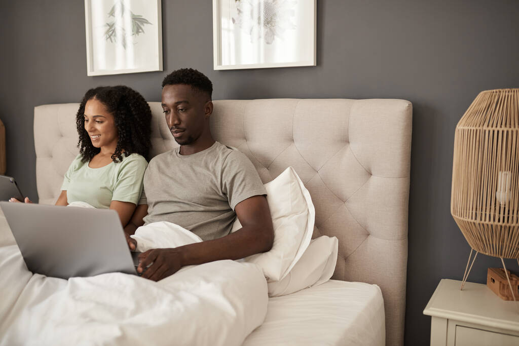 jong multiraciaal paar zitten in hun bed thuis in de ochtend en met behulp van een laptop en digitale tablet - Foto, afbeelding