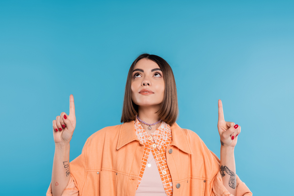 summer fashion, brunette young woman with short hair and piercing in nose and tattoos posing in casual outfit on blue background, everyday makeup, orange shirt, generation z, showing and pointing up  - Photo, Image