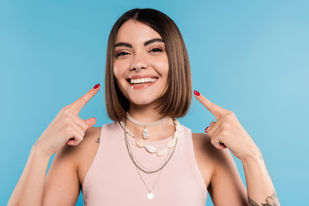 excitement, tattooed young woman with short brunette hair in tank top smiling and pointing at her cheeks on blue background, casual attire, gen z fashion, happiness, joyful  - Photo, Image