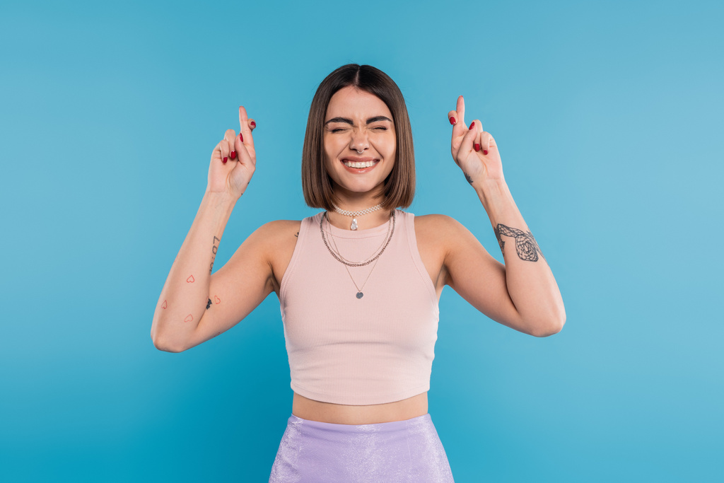fingers crossed, tattooed young woman with short brunette hair in tank top making wish on blue background, casual attire, gen z fashion, happiness, smiling with closed eyes  - Photo, Image