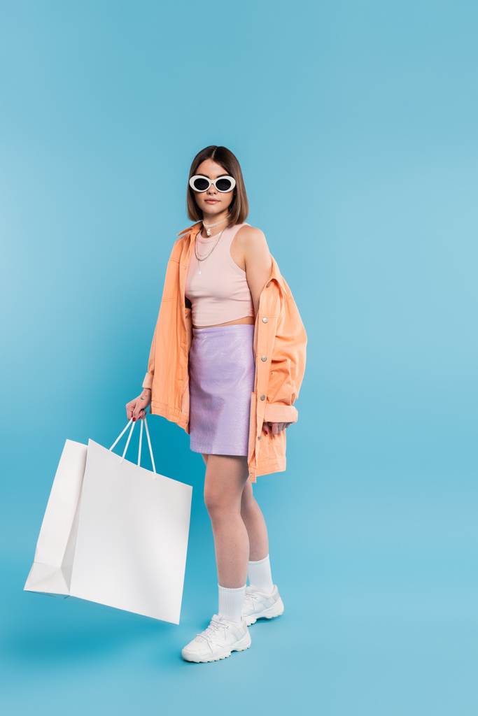 летние шоппинг, брюнетка молодая женщина в майке, юбка, солнцезащитные очки и оранжевая рубашка позирует с сумкой для покупок на голубом фоне, случайный наряд, стильная поза, ген z, современная мода  - Фото, изображение