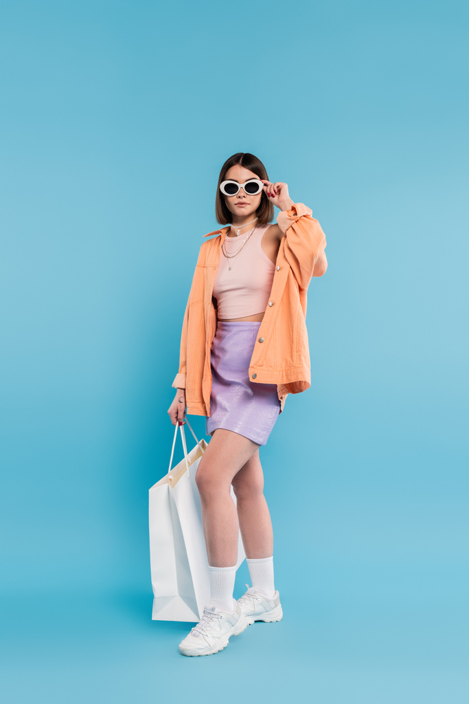 módní nakupování, brunetka mladá žena v tílku, sukně, sluneční brýle a oranžová košile pózující s nákupní taškou na modrém pozadí, ležérní oblečení, stylové pózování, gen z, moderní móda  - Fotografie, Obrázek