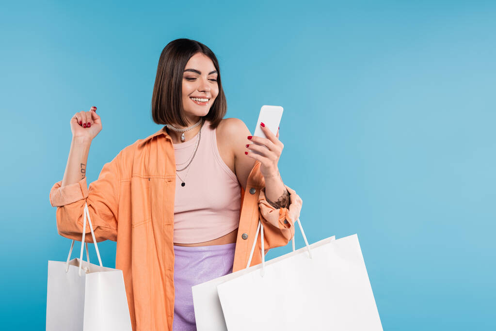 positivity, shopping spree, cheerful young woman in trendy outfit holding shopping bags and using smartphone on blue background, casual attire, stylish, generation z, modern fashion  - Photo, Image