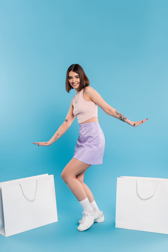 positivity, summer fashion, shopping spree, tattooed young woman with nose piercing posing in trendy outfit near shopping bags on blue background, casual attire, generation z, modern fashion  - Photo, Image