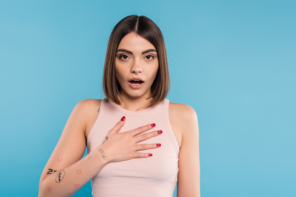 shocked woman with hand on chest, stylish model with tattoos and nose piercing looking at camera on blue background, emotional, opened mouth, generation z, summer fashion  - Photo, Image