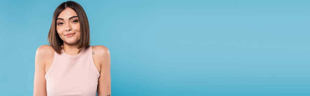 not knowing, positive young woman with tattoos and nose piercing standing in tank top on blue background, looking at camera, confused, pretty face, generation z, summer outfit, banner  - Photo, Image