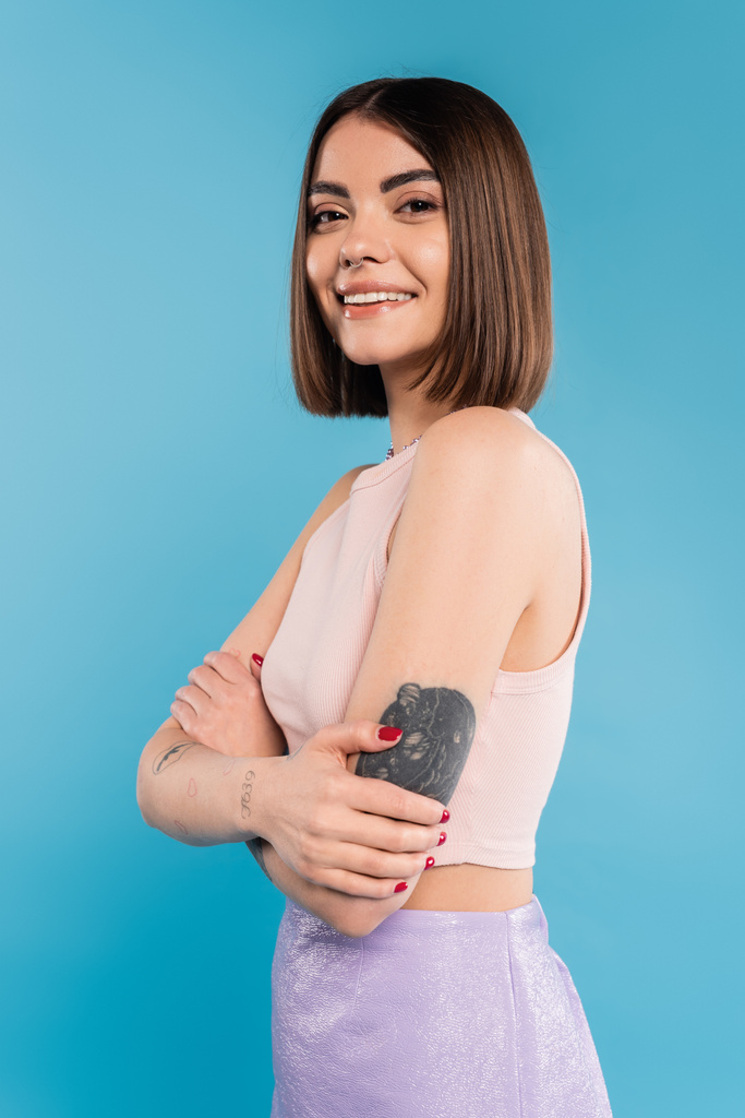Katlanmış eller, kısa saçlı ve dövmeli neşeli genç bir kadın mavi arka planda kameraya bakıyor, burun piercingli, gençlik, Z kuşağı, yaz modası  - Fotoğraf, Görsel
