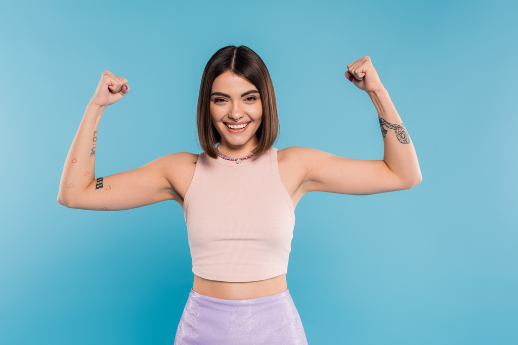 women power, cheerful young woman with short hair, tattoos and nose piercing showing muscles on blue background, generation z, displeased, casual attire, strength  - Photo, Image