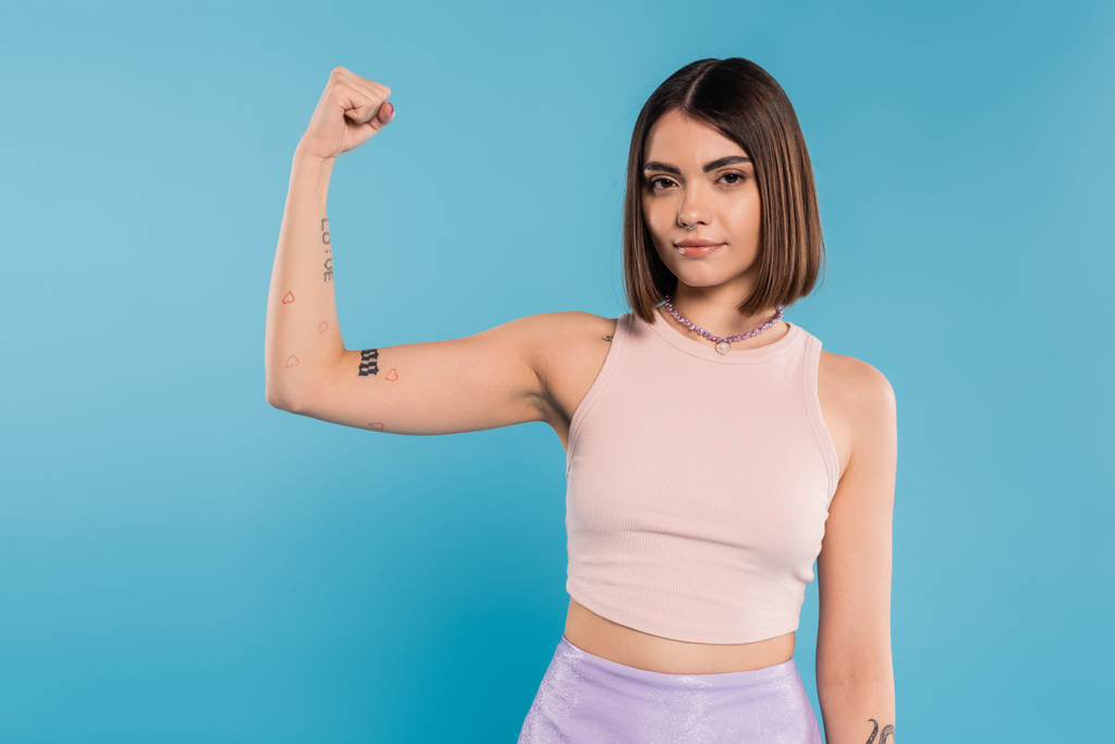 female power, brunette young woman with short hair, tattoos and nose piercing showing muscle on blue background, generation z, displeased, casual attire, strength  - Photo, Image