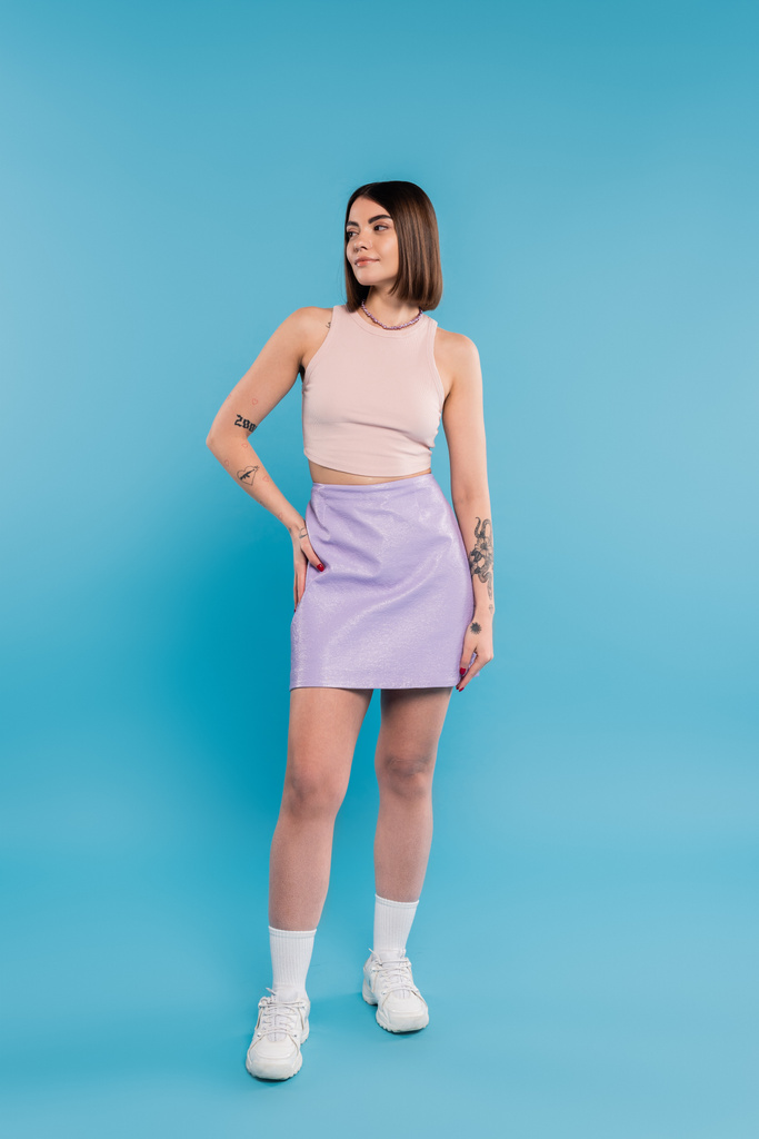 summer trends, brunette young woman with short hair in tank top, skirt and white sneakers posing on blue background, casual attire, gen z fashion, personal style, nose piercing, hand on hip  - Photo, Image