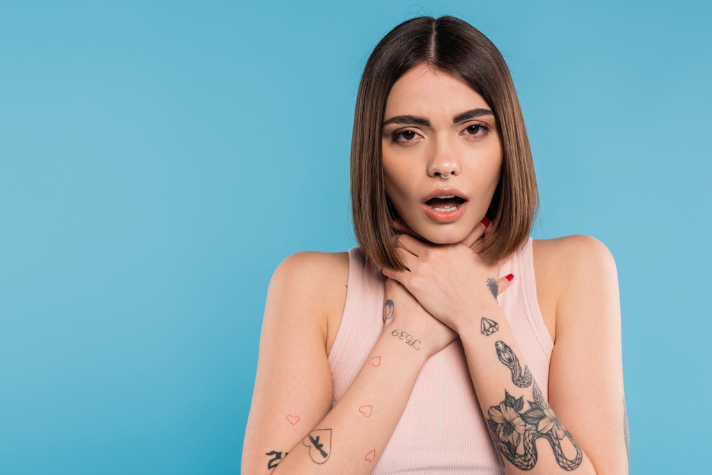 brunette young woman with short hair holding hands near neck and standing with opened mouth while choking on blue background, casual attire, gen z fashion, personal style, nose piercing  - Photo, Image