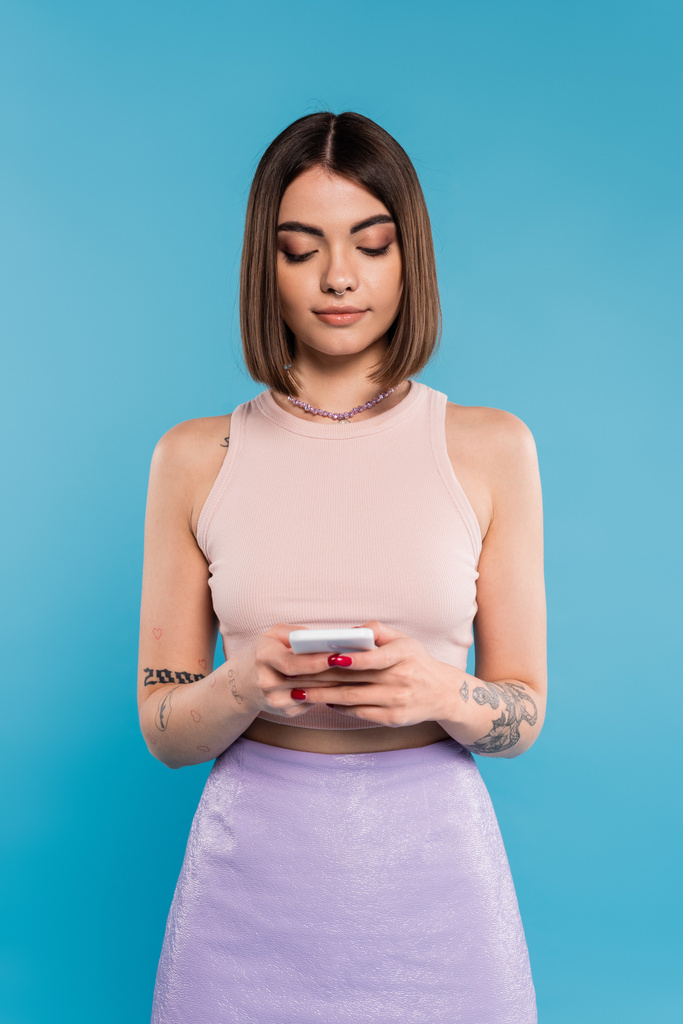 mujer mensajes de texto en el teléfono inteligente, pelo corto, tatuajes y piercing en la nariz usando el teléfono móvil sobre fondo azul, atuendo casual, moda gen z, estilo personal, maquillaje diario  - Foto, imagen