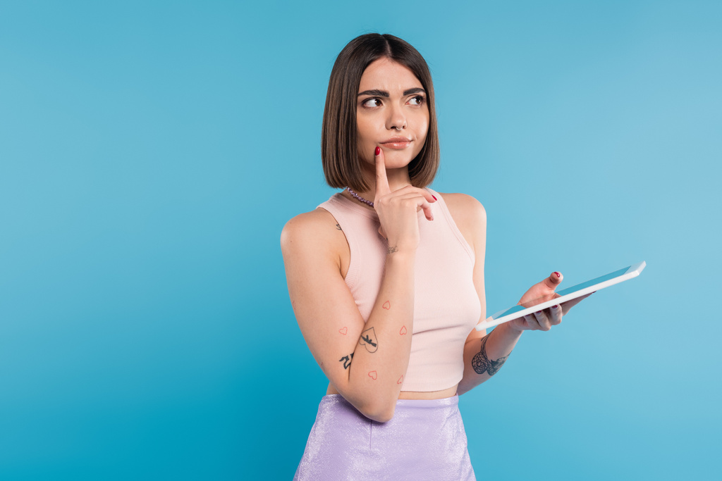 brunette young woman with short hair, tattoos and nose piercing digital tablet laptop on blue background, generation z, summer trends, attractive, social media influencers, tablet user, pensive  - Photo, Image
