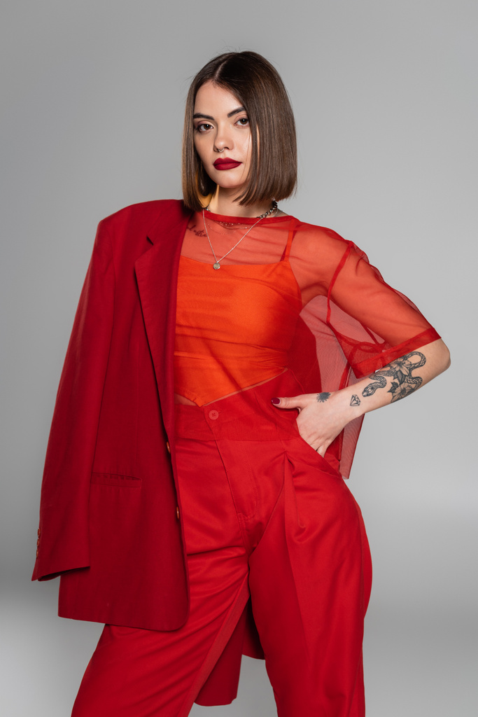 stylish suit, bold makeup, young tattooed woman with short hair holding red blazer on grey background, generation z, trendy outfit,  professional attire, executive style  - Photo, Image