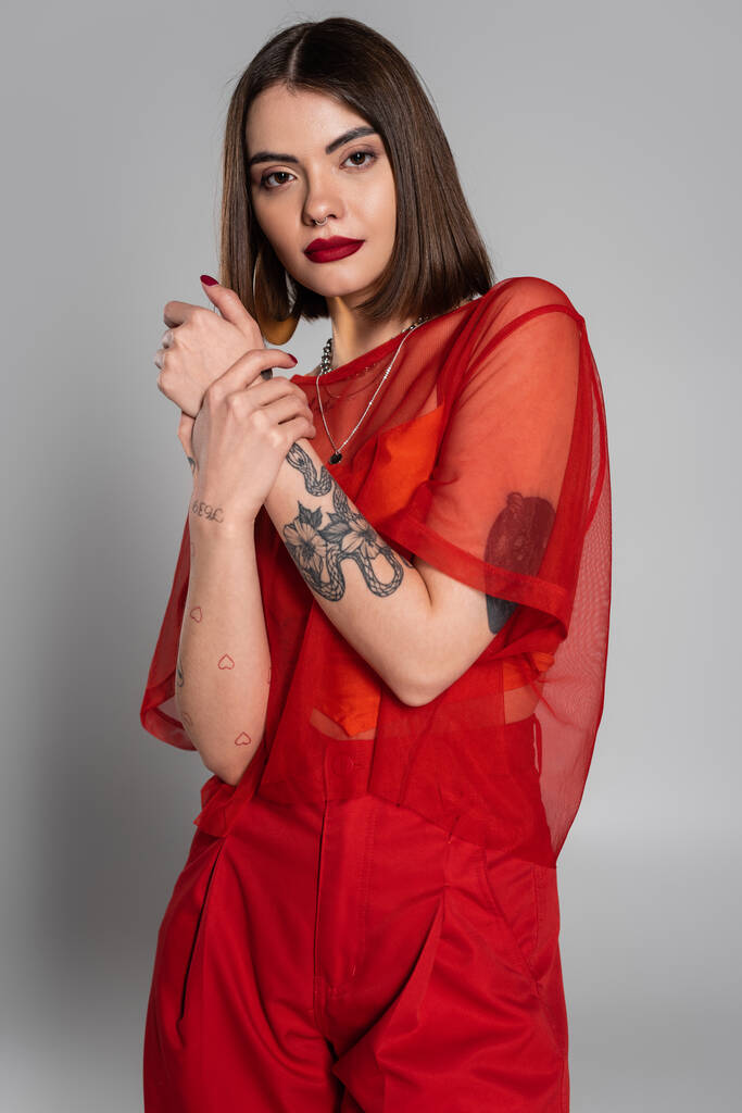 divat, piros ruha, tetovált és fiatal nő rövid haj és orr piercing pózol átlátszó blúz és nadrág szürke háttér, modern stílus, generáció z, divat - Fotó, kép