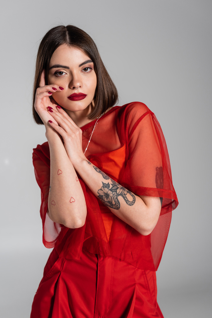 модель, красный наряд, татуированная молодая женщина с короткими волосами и пирсингом носа позирует в красной прозрачной блузке на сером фоне, современный стиль, поколение z, мода - Фото, изображение